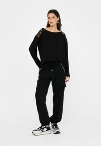 Sort, langærmet top med blonder ved skuldrene og løs pasform, kombineret med sorte joggers med sidelommer og snøre i taljen.