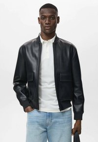 Chaqueta bomber de cuero negro con cremallera, dos bolsillos frontales y puños acanalados, llevada sobre una camiseta polo blanca y jeans azul claro.
