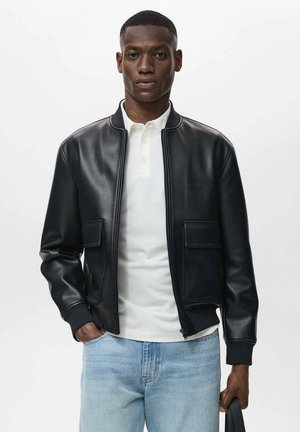 Chaqueta bomber de cuero negro con cremallera, dos bolsillos frontales y puños acanalados, llevada sobre una camiseta polo blanca y jeans azul claro.
