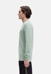 Suéter de punto verde claro con cuello redondo, puños acanalados, que presenta un pequeño logo en el pecho, combinado con pantalones oscuros.