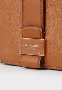Dettaglio di una borsa a mano in pelle marrone con una placca in tono oro che riporta la scritta "kate spade NEW YORK", con una finitura testurizzata e cuciture visibili.