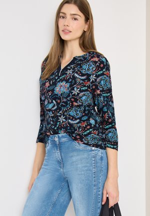 Femme portant un chemisier à manches longues à fleurs foncées et un jean bleu clair, debout contre un mur blanc, tenant un sac noir dans sa main droite.