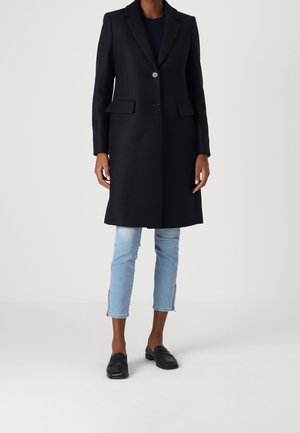 Persoon draagt een zwarte knielange jas met knopen, lichtblauwe cropped jeans met ritssluitingen aan de zijkant, en zwarte loafers, staand op een witte vloer.