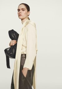 Lichtcrème lange mouw top, gelaagd met een beige sjaal, gecombineerd met donkerbruine leren broeken. Houdt een zwarte leren tas met een hengsel vast.