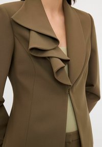 Olivově zelený blazer s velkým řaseným límcem, texturovanou látkou, zapínáním na jeden knoflík a přiléhavým střihem, nošený přes světle zelený top.
