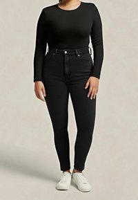 Personne portant un haut noir ajusté à manches longues, un jean skinny noir taille haute et des baskets blanches à lacets, debout sur un fond uni.