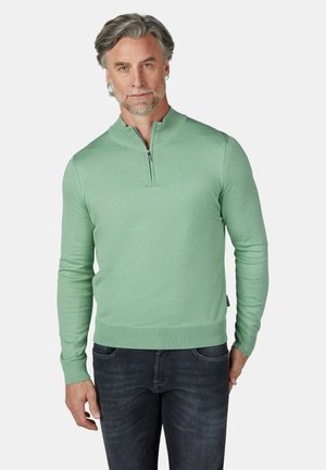 Man met grijs haar en baard, gekleed in een lichtgroene lange mouwen trui met rits en donkerblauwe spijkerbroek, staand met één hand in de zak.