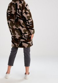Lang, camoufleret jakke i blød, fuzzy stof med nuancer af grøn, brun og beige; sat sammen med grå cropped jeans og hvide sneakers.