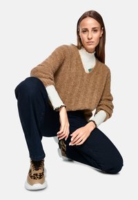 Brauner, flauschiger Strickpulli mit V-Ausschnitt, kombiniert mit dunklen Jeans. Dazu kommen gemusterte Sneaker und ein weißes Langarmshirt darunter.