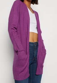 Fialový pletený cardigan s uvolněným střihem, otevřeným předním dílem, dlouhými rukávy a dvěma předními kapsami. Měkká textura s žebrovanými manžetami a lemem.