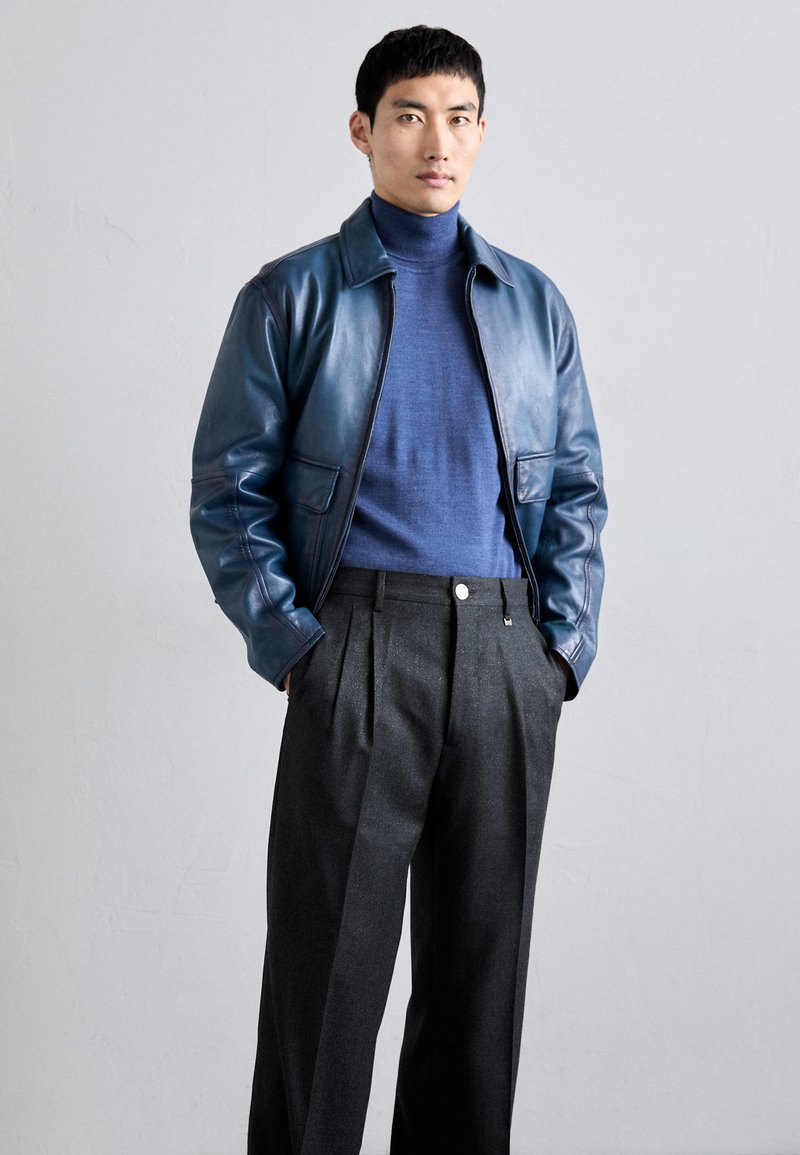 Giubbotto bomber in pelle di colore blu scuro, abbinato a un dolcevita blu e pantaloni grigi plissettati, che mettono in risalto una texture liscia e un design contemporaneo.