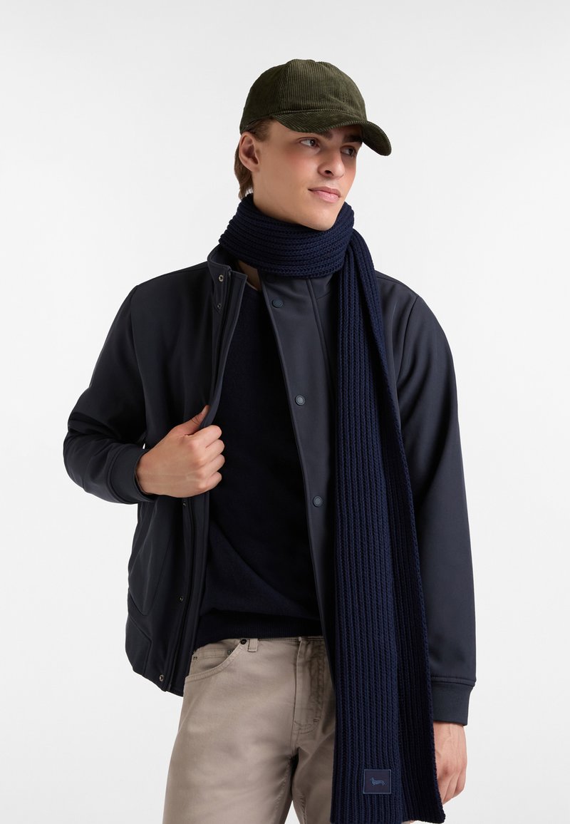 Giovane uomo con un cappello verde oliva, giacca blu navy e sciarpa, e pantaloni beigi che guarda di lato su uno sfondo bianco.