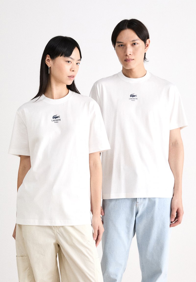 Hvit t-skjorte med kort erme og Lacoste-logo i marineblått. Laget av bomull, med en avslappet passform og rund hals. Brukt med lyse denim og beige bukser.