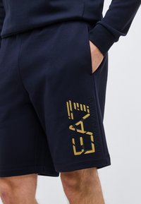 Άνδρας που φοράει ναυτικού μπλε σορτς με χρυσό λογότυπο Emporio Armani, το χέρι στην πλαϊνή τσέπη, ορατό μέρος από το αντιστοιχούμνο μακρυμάνικο μπλε τοπ.