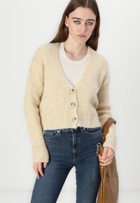 Cardigan en tricot beige avec un décolleté en V, trois grands boutons, des poignets côtelés et une coupe décontractée, associé à un jean bleu foncé.