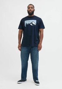 Erwachsener Mann mit Bart, der ein dunkelblaues T-Shirt mit einem Berg- und Baum-Graphic trägt, dazu blaue Jeans und schwarze Sneakers, vor einem weißen Hintergrund stehend.