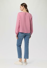 Rosa langärmlige Bluse mit einer Rückenöffnung, kombiniert mit hellblauen Jeans und beigen Sneakern mit metallischen Akzenten.