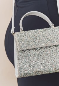 Bolso de mano plateado brillante adornado con cristales multicolores, con una forma estructurada, asa superior y correa de hombro desmontable.