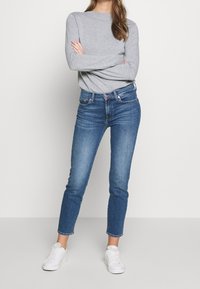 Šedá dlouhá rukávová košile v kombinaci s modrými skinny džíny. Džíny mají klasický design s pěti kapsami a jsou obuty s bílými teniskami.