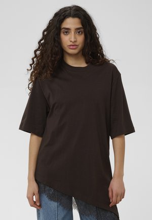 Lokkis juustega naine, kes kannab lahtist musta asümmeetrist T-särki pitsilise servaga teksapükste kohal, seisab ühevärvilise tausta ees.