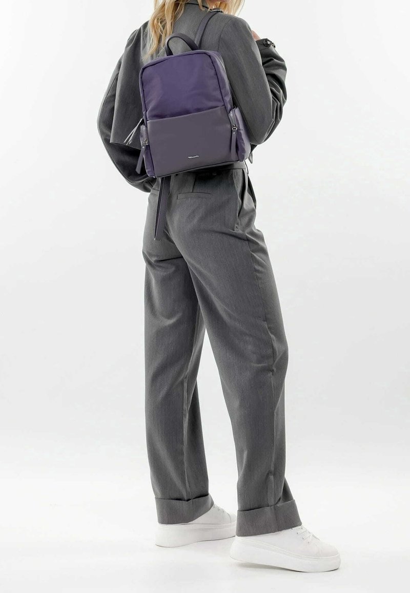 Sac à dos en nylon violet de forme rectangulaire, avec des accents zippés et des bretelles réglables, associé à un pantalon gris et des baskets blanches.