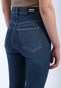 Blue denim jeans with a high waist, featuring a black logo patch at the back, two back pockets, and contrast stitching detailing.