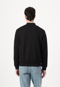 Svart bomberjakke med teksturert mønster, ribbestrikkede mansjetter og kant, samt en klassisk krage, sammen med lyseblå denimjeans.