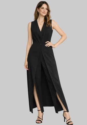 Kobieta ubrana w czarną, długą kamizelkę bez rękawów z przednimi rozcięciami oraz czarne sandały na wysokim obcasie, stojąca z ręką na biodrze na szarym tle.