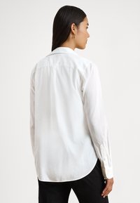 Camisa branca de mangas compridas feita de tecido leve, com bainha arredondada, punhos abotoados e colarinho clássico.