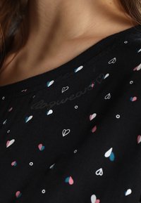 Zbliżenie czarnej bluzki z małym wzorem serc i kółek w kolorze białym, różowym i niebieskim blisko szyi osoby oraz jej włosów.