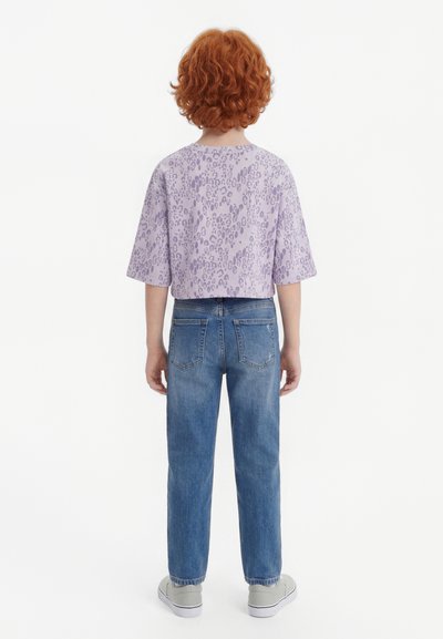 Kind mit lockigem rotem Haar, das ein lavendelfarbenes Leopardenmuster-Shirt, blaue Jeans und graue Turnschuhe trägt, steht mit dem Rücken zur Kamera vor weißem Hintergrund.