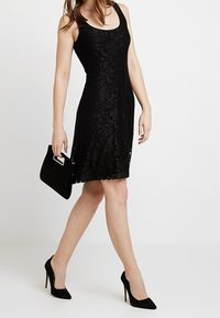 Robe en dentelle noire avec une silhouette ajustée, un col rond et un design sans manches. Associée à une pochette noire et des chaussures à talons hauts.