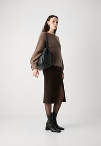 Pulover din lână maro cu mâneci tip balon, fustă maro cu rimel și fante, și botine negre cu toc. Geantă neagră pe umăr. Fundal minimalist.