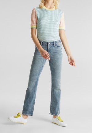 Lichtblauwe, textuur gebreide top met korte pastel mouwen, gecombineerd met lichtgewassen spijkerbroek en witte sneakers met gele accenten.