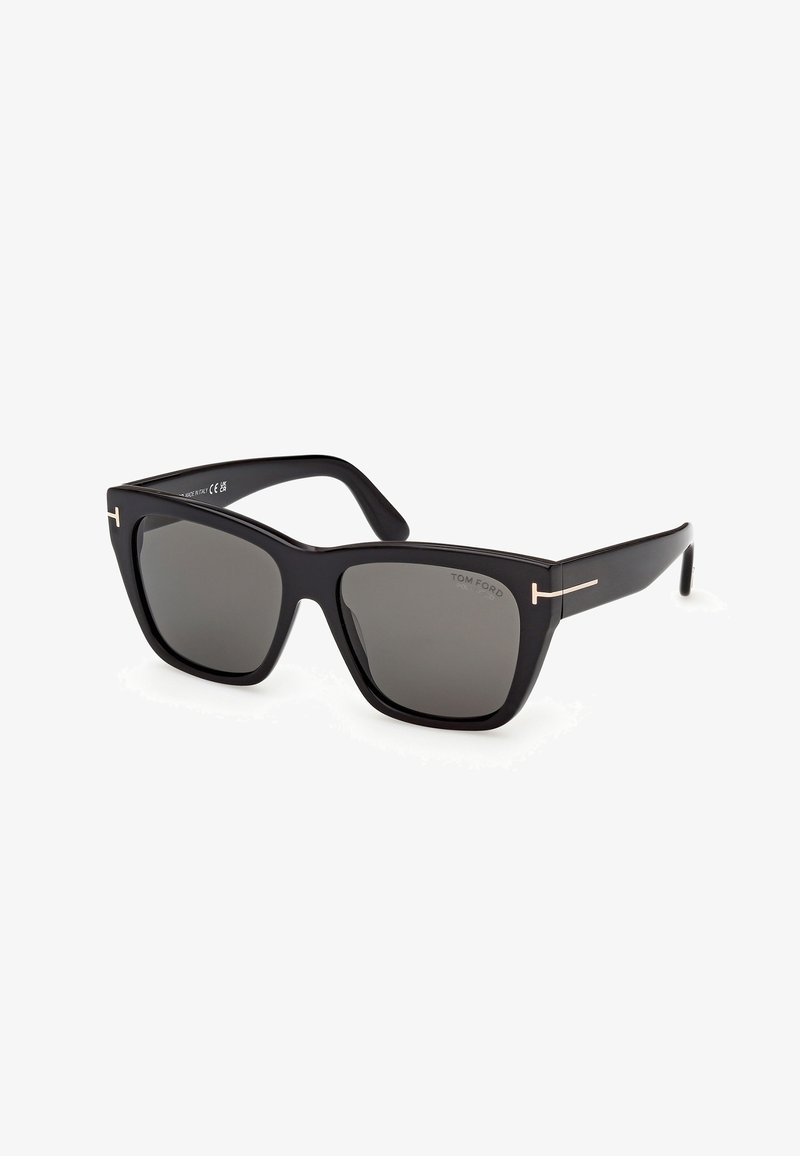 Lunettes de soleil carrées noires surdimensionnées avec des verres foncés et des accents en forme de T fins dorés sur les branches.