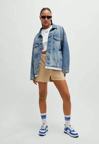 Casaco de ganga em azul claro, top cropped branco, calções beges e sapatilhas com pormenores azuis. Os acessórios incluem óculos de sol pretos e meias estampadas.