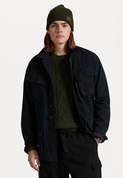 Veste bleu marine foncé avec deux grandes poches poitrine, superposée à un pull vert texturé. Portée avec un bonnet vert et un pantalon noir.