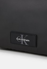 Black Calvin Klein bag with a smooth, matte texture. Features a rectangular logo patch with embossed lettering. Minimalist design.