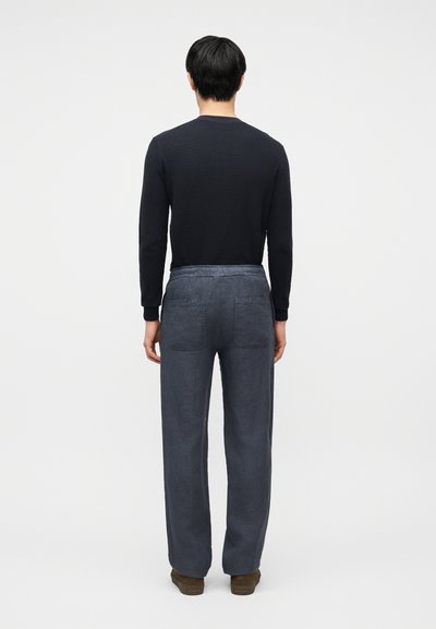 Άνδρας που στέκεται με την πλάτη προς τα εμπρός, φοράει μαύρο πουκάμισο με μακριά μανίκια, χαλαρά σκούρα γκρι παντελόνια με πίσω τσέπες και καφέ παπούτσια σε απλό φόντο.