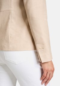 Beżowa lniana marynarka o gładkiej fakturze, z pojedynczym guzikiem na mankietach, zestawiona z białymi dżinsami.