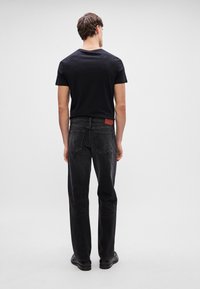 Tricou negru, material moale. Blugi negri din denim, croi drept, textură uzată. Patch de marcă din piele marron la talie. Pantofi negri.