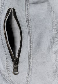 Detail einer grauen Lederjacke mit einer Reißverschlusstasche, die einen metallenen Reißverschluss und kontrastierende Nähte entlang der Kanten aufweist.