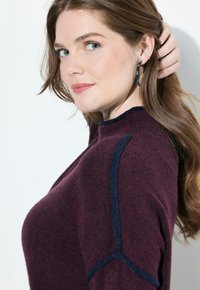 Maglione bordeaux con accenti blu scuro sulle spalle, texture morbida e scollatura rotonda. Indossato con grandi orecchini a cerchio.