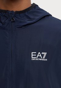 Mørkeblå zip-up jakke med hætte, med en sort krave og et hvidt EA7 Emporio Armani logo på venstre side. Glat, letvægtsstof.