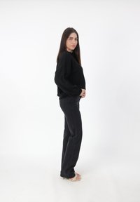 Maglione nero lavorato a maglia con maniche lunghe abbinato a jeans neri a vita alta. Il modello indossa tacchi beige su uno sfondo bianco.