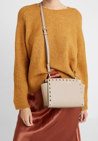 Brązowa torebka z pudełkowymi ćwiekami, akcentem z logo i regulowanym paskiem. Noszona z pomarańczowym swetrem w prążki i satynową spódnicą w kolorze rdzy.