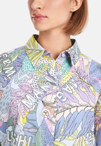 Farbige Hemdbluse mit einer Mischung aus Pastellmustern, einschließlich Blättern und abstrakten Formen, mit einem klassischen Kragen und einer glatten Stofftextur.