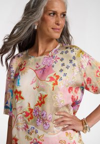 Mujer con cabello largo y canoso, con pendientes plateados y blusa con estampado floral, posando con la mano en la cadera frente a un fondo liso.