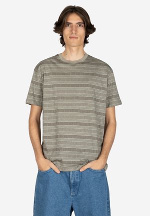 Junger Mann mit schulterlangem braunem Haar, der ein graues gemustertes Kurzarmhemd und blaue Jeansshorts trägt, Hände in den Taschen, neutraler Hintergrund.