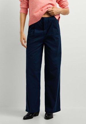 Person trägt marineblaue weite Hosen, schwarze Absatzschuhe und einen rosa Pullover, der hochgezogen ist, um die Taille der Hose sichtbar zu machen.
